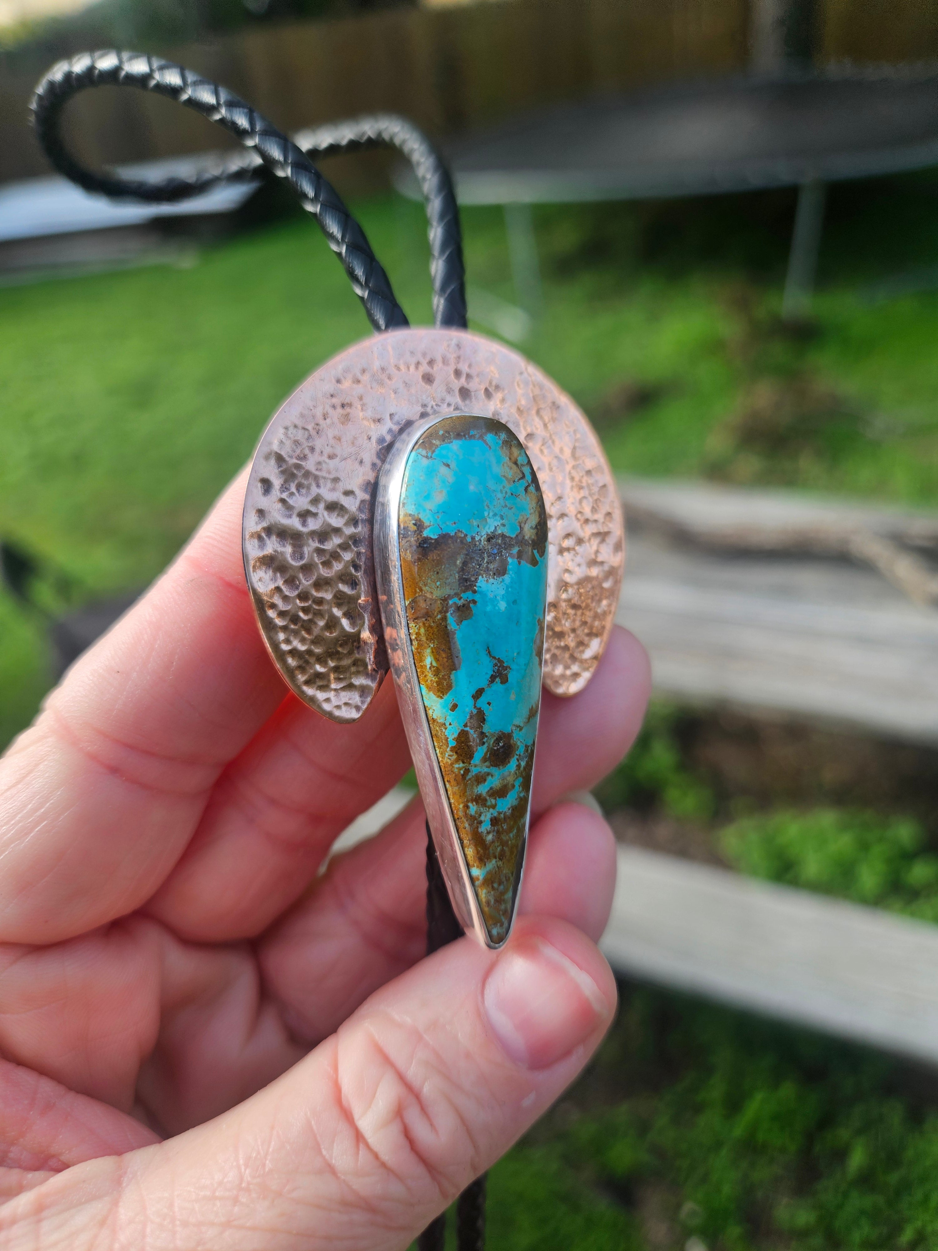 A handmade turquoise and copper pendant bolo featuring a hammered copper disc and a teardrop-shaped turquoise stone with copper matrix, on a black cord.